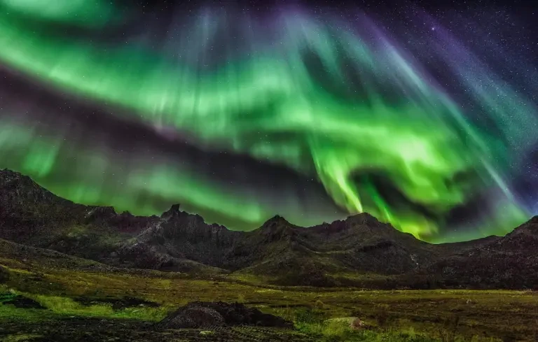 Aurora hijau melengkung di langit malam di atas pegunungan bersalju yang terpantul di air.