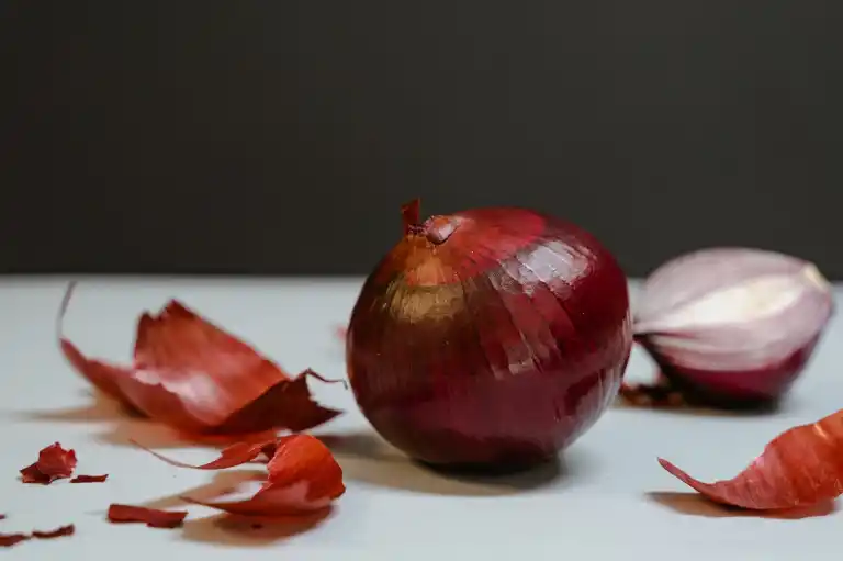 Satu bawang merah besar berkulit ungu tua di atas meja putih, dengan serpihan kulitnya berserakan dan satu potongan di belakangnya.
