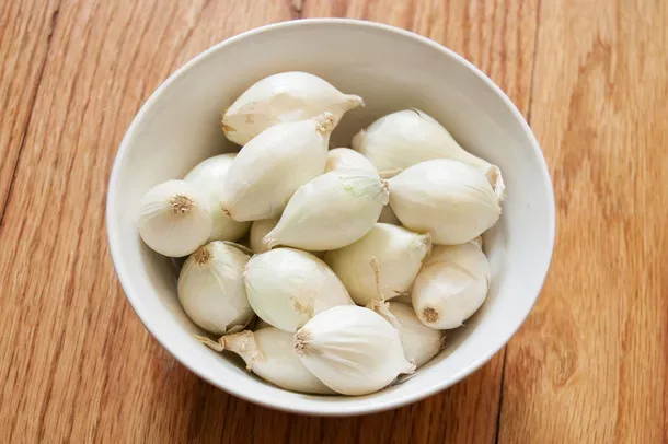 Semangkuk bawang mutiara putih kecil-kecil berbentuk bulat rapi, di atas meja kayu.